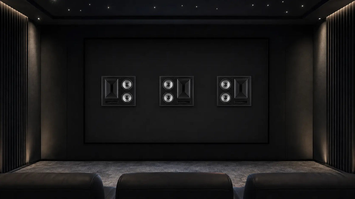 A dark home theater or audio room with three black speaker cabinets mounted on the back wall above a carpeted floor, with rows of dark seat backs visible in the foreground.